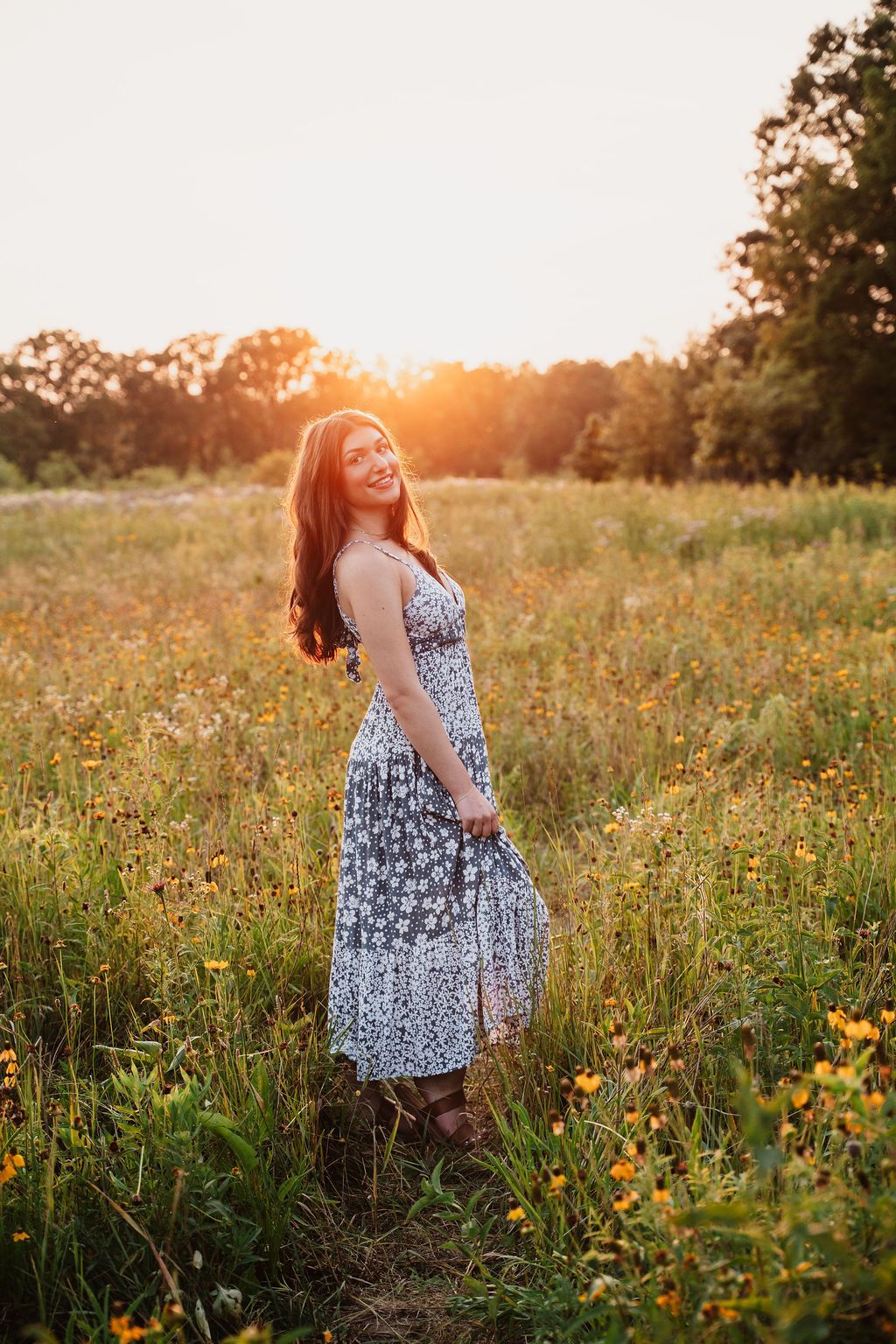 Young woman in floral sundress smiling in golden sunset meadow with wildflowers and trees in background.