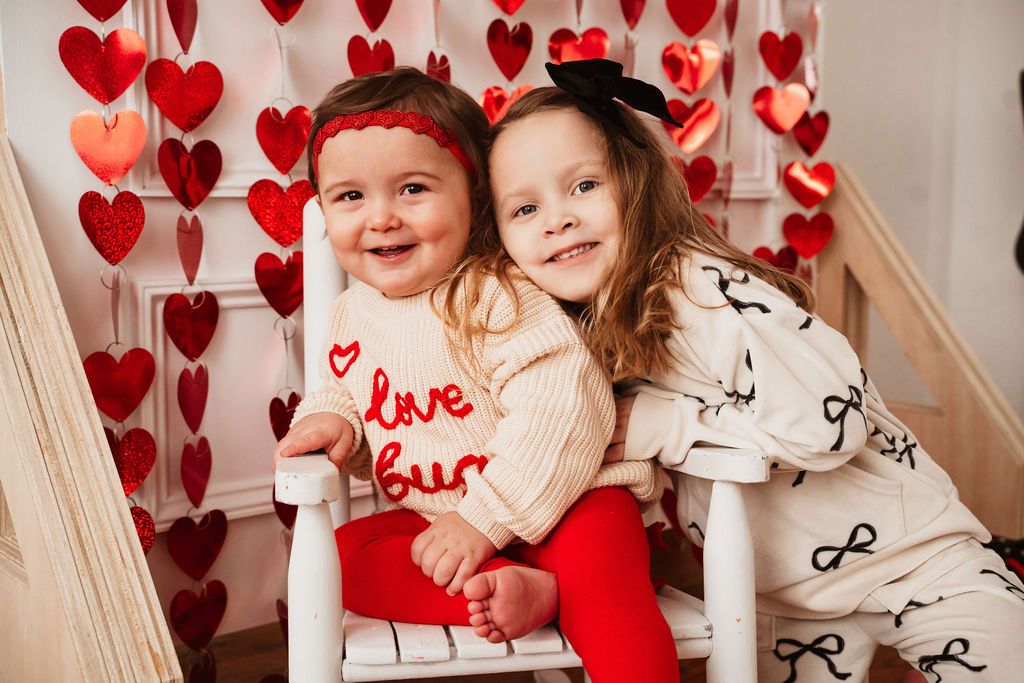 Two young sisters pose together in Valentine's Day outfits with red heart decorations in the background.