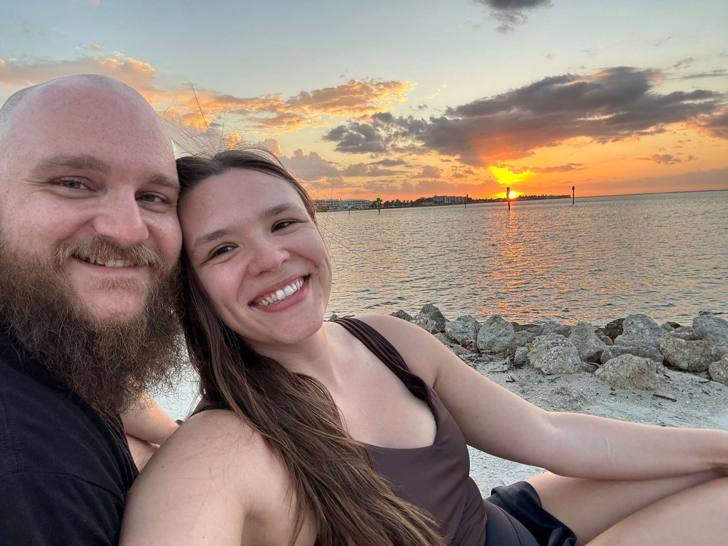 Smiling couple taking a selfie at sunset by rocky shoreline with vibrant orange and pink sky over water.