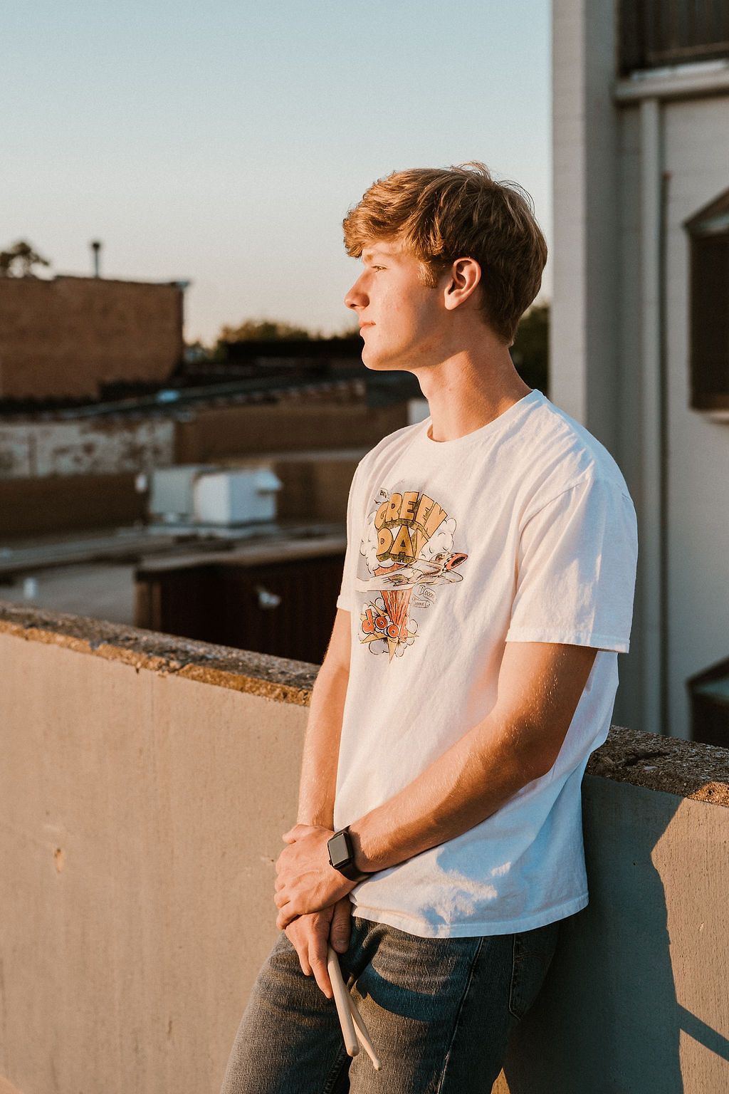 Young man in white graphic tee and jeans stands on rooftop at golden hour, gazing into the distance.