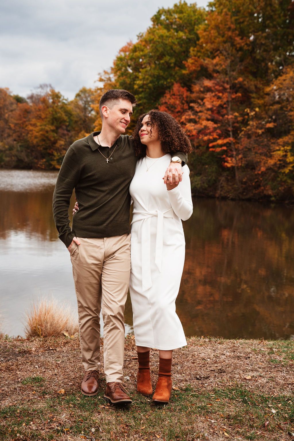 Couple embracing by a lakeside in autumn, surrounded by colorful fall foliage, woman in white dress, man in green sweater.