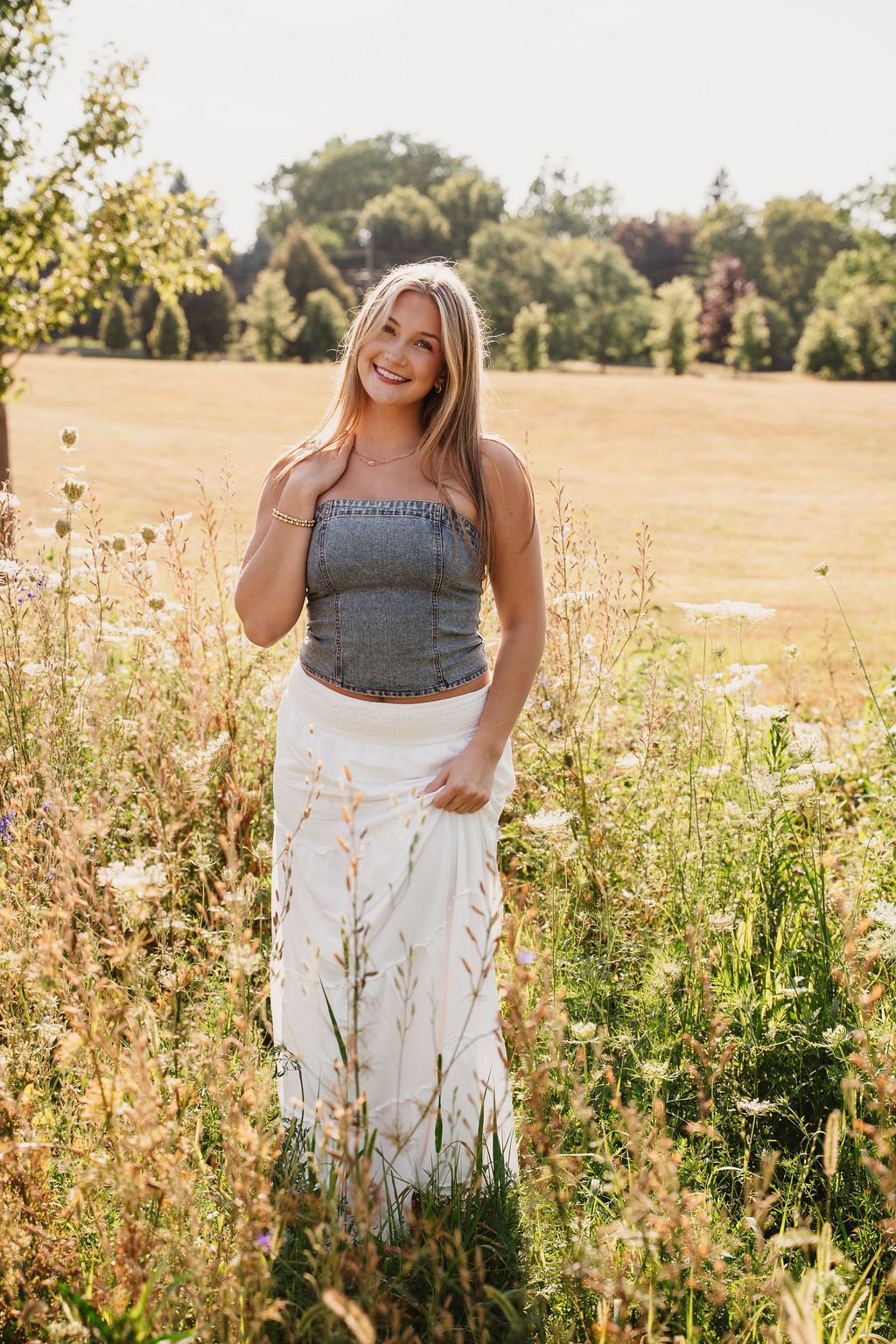 Smiling blonde woman in gray crop top and white maxi skirt standing in a sunny wildflower field.