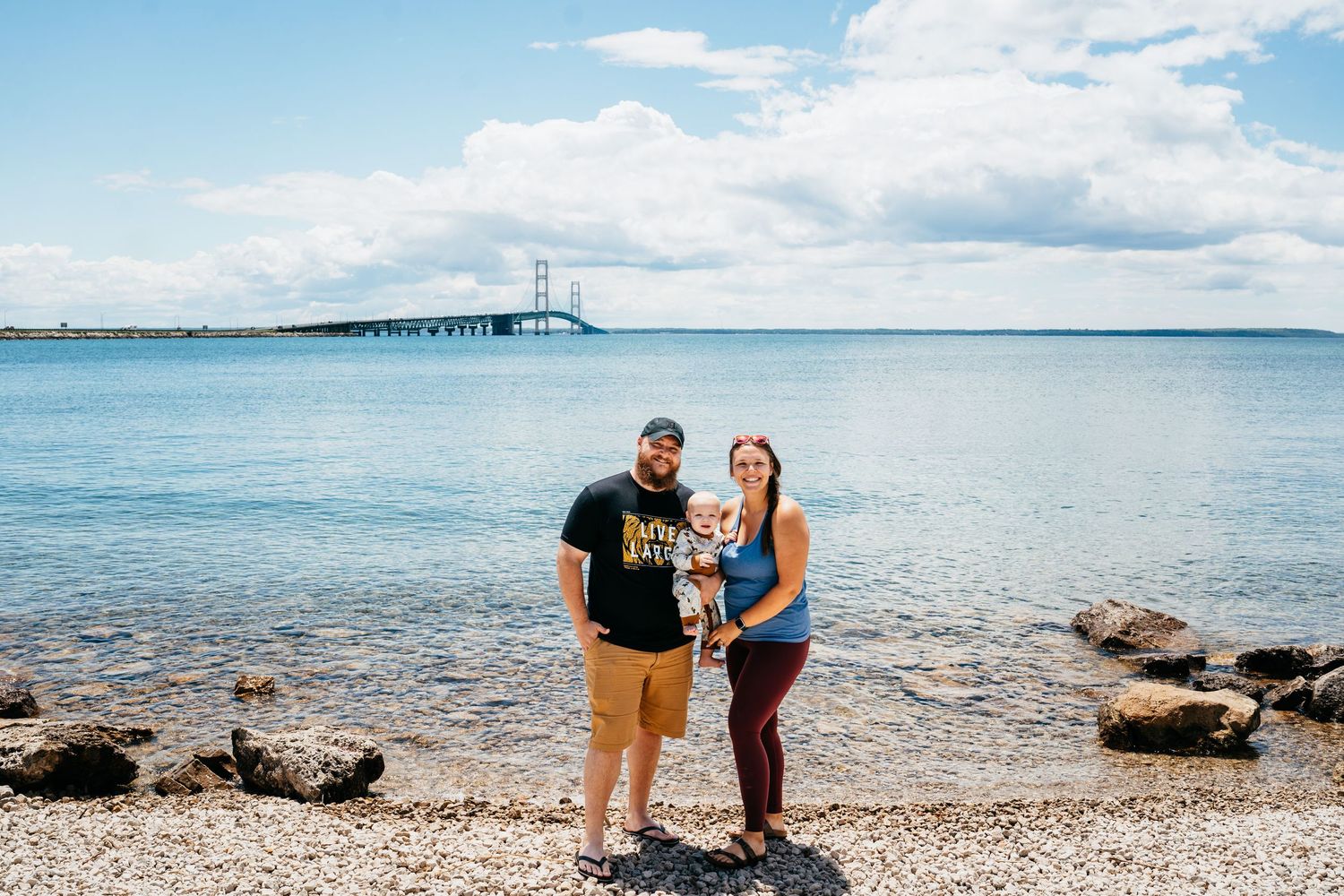 Family of three posing on rocky shore with Mackinac Bridge spanning blue waters under cloudy sky.