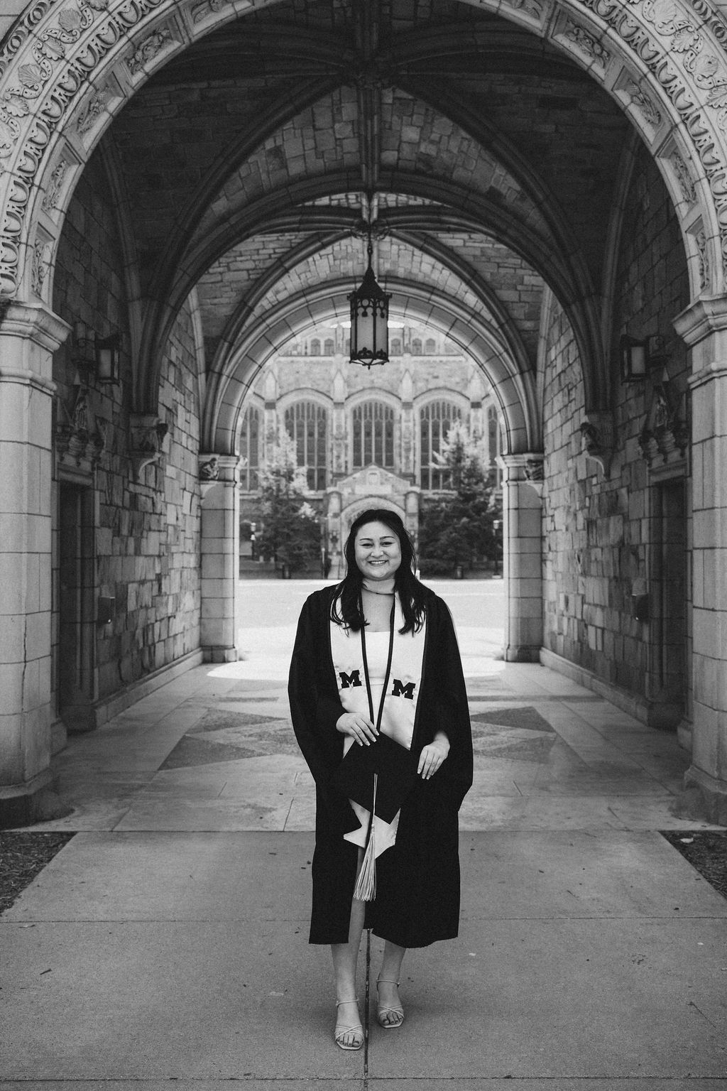 Black and white photo of graduate in cap and gown smiling under gothic stone arch at U of M.