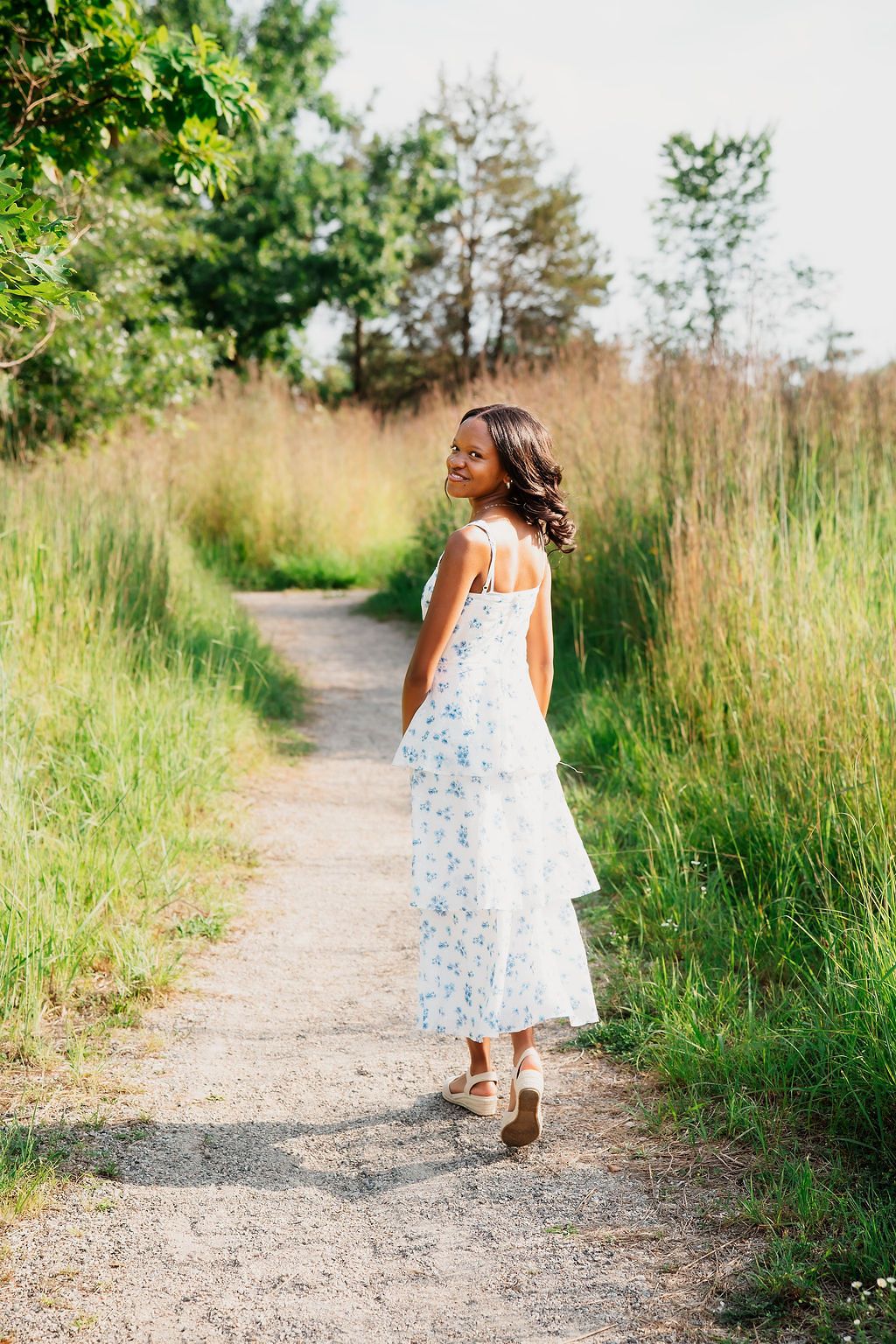 A child in a white summer dress walks along a winding dirt path surrounded by tall grass and greenery.
