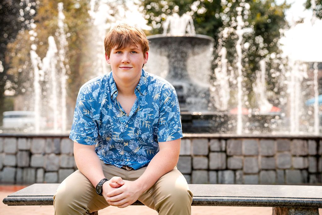 Young man in blue floral shirt sits on bench before a fountain, smiling outdoors in natural light.