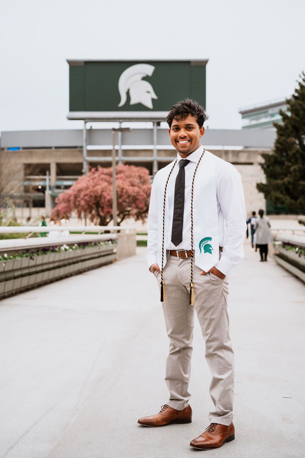A person in a white shirt and khaki pants poses for graduation photos on a concrete walkway at Michigan State University.