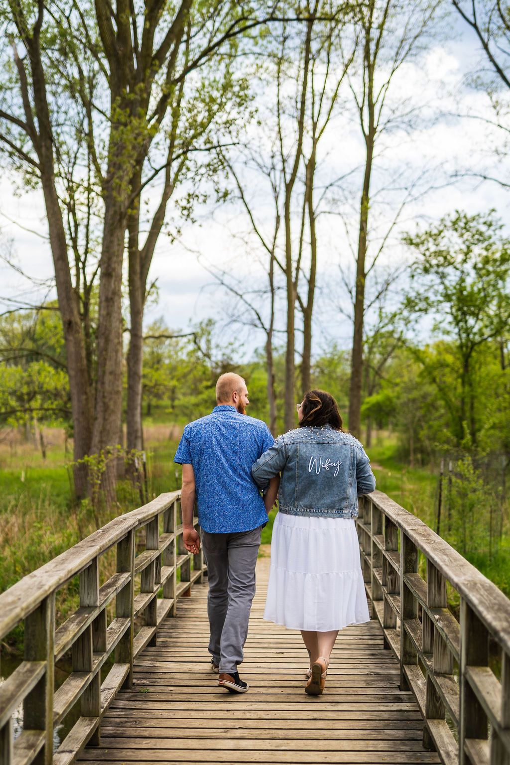 Two people walk together holding hands on a wooden boardwalk through a scenic wooded nature area on a spring day.