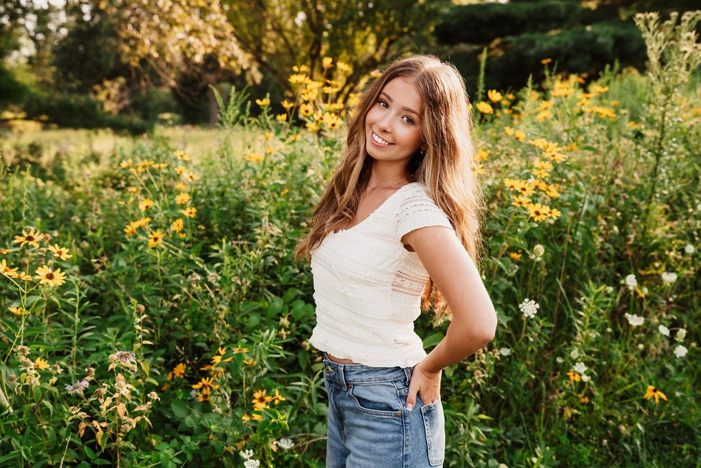 A person in a white top and jeans poses in a vibrant field of yellow wildflowers during golden hour.