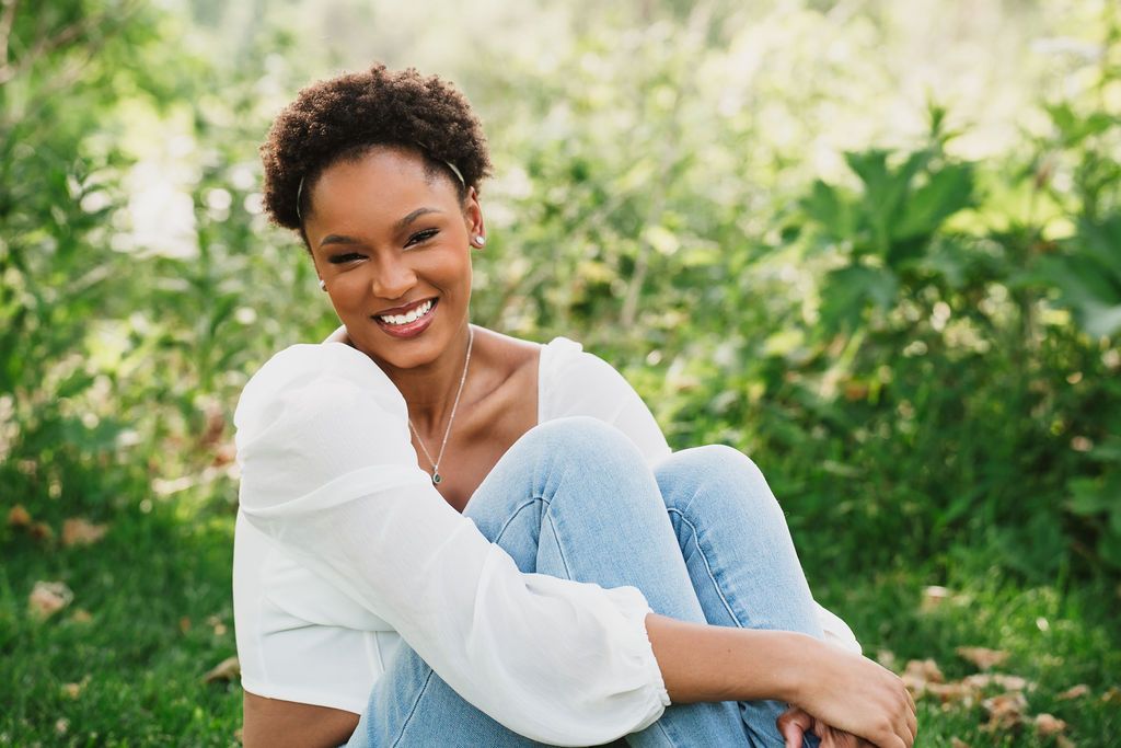 Person in casual attire smiling and enjoying a sunny day in a lush green outdoor setting.