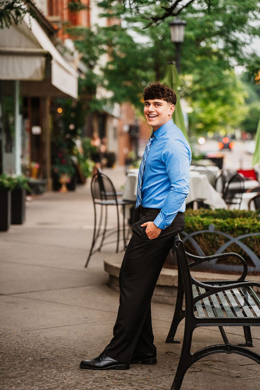 Young man in blue shirt and black pants smiling on urban sidewalk near bench and outdoor cafe.