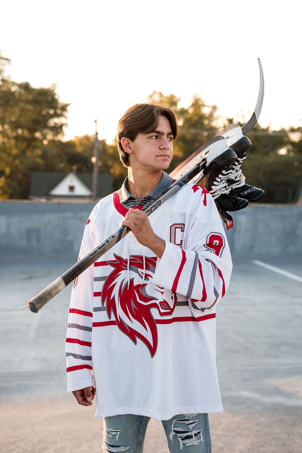 Teen hockey player in white jersey carrying stick and gloves on rooftop at sunset.