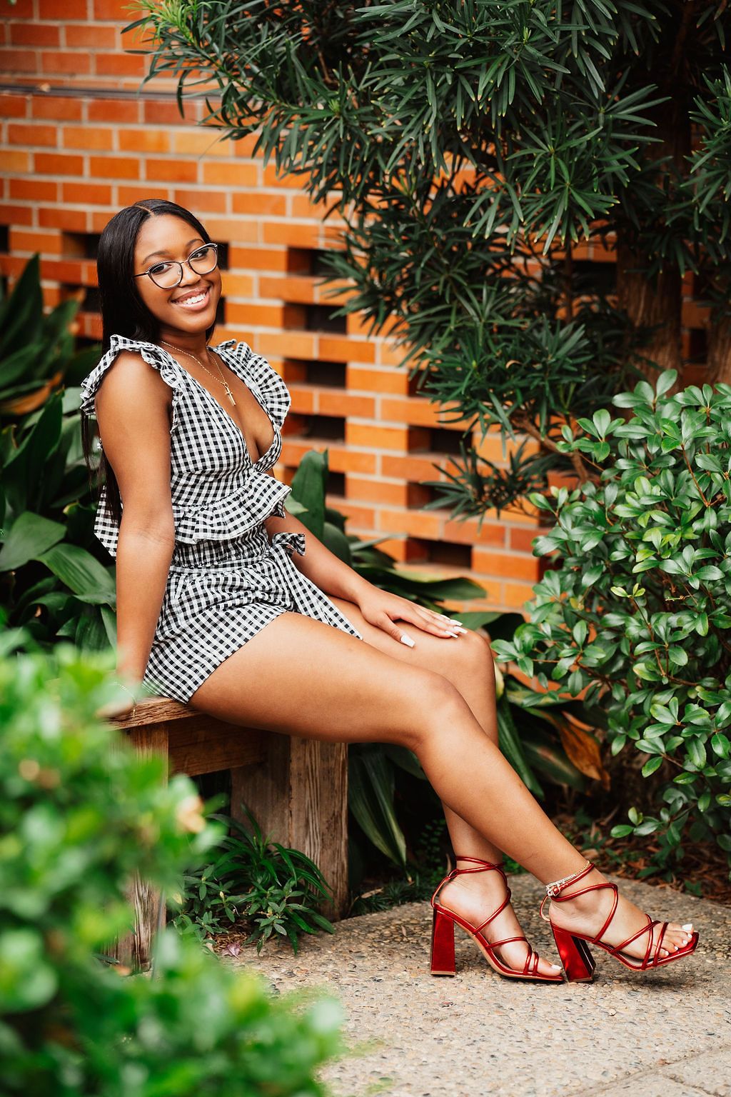 Red sandals and a checkered dress complement a rustic brick wall and garden backdrop.