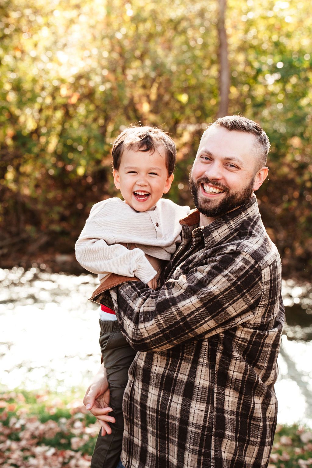 Smiling father holding laughing toddler outdoors in autumn park setting with golden foliage background.
