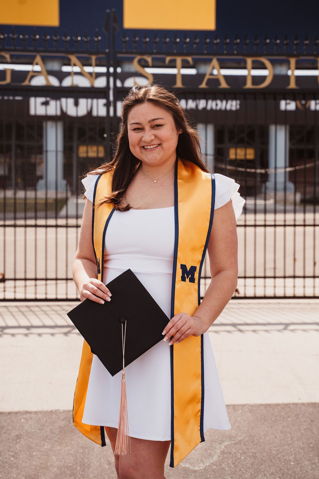 Smiling female graduate in white dress with gold Michigan stole holds cap outside Michigan Stadium.
