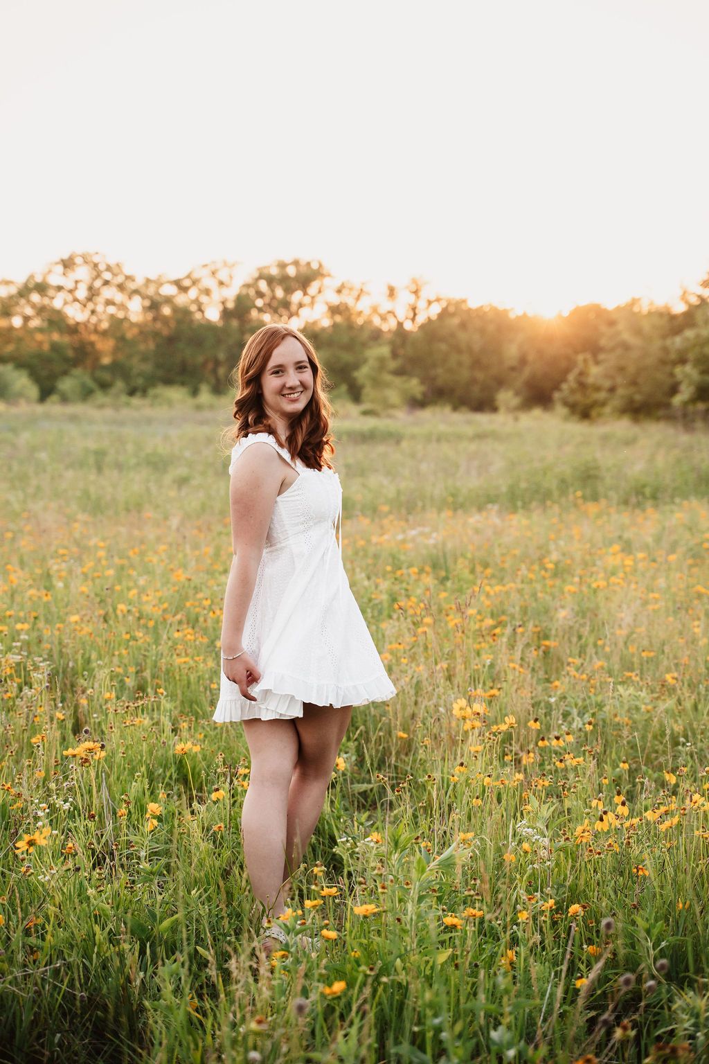 Young woman in white dress standing in golden hour wildflower field, smiling warmly at camera.