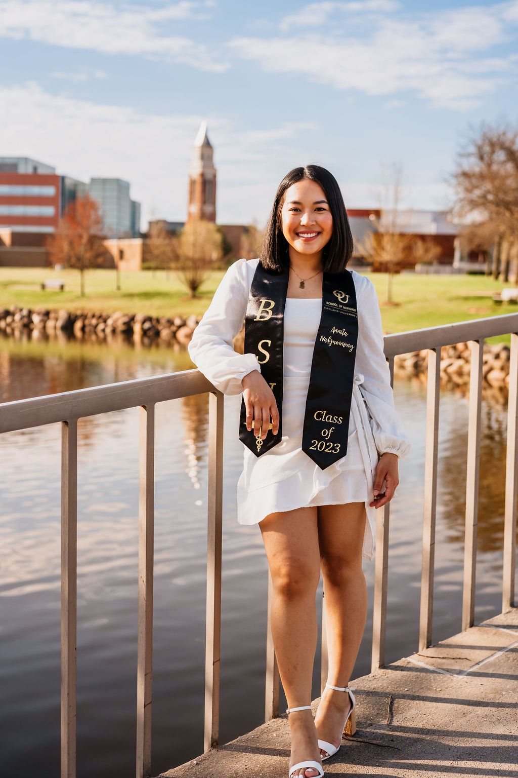 A graduate in a white dress and decorated stole poses on a bridge with a church spire and campus buildings in the background.