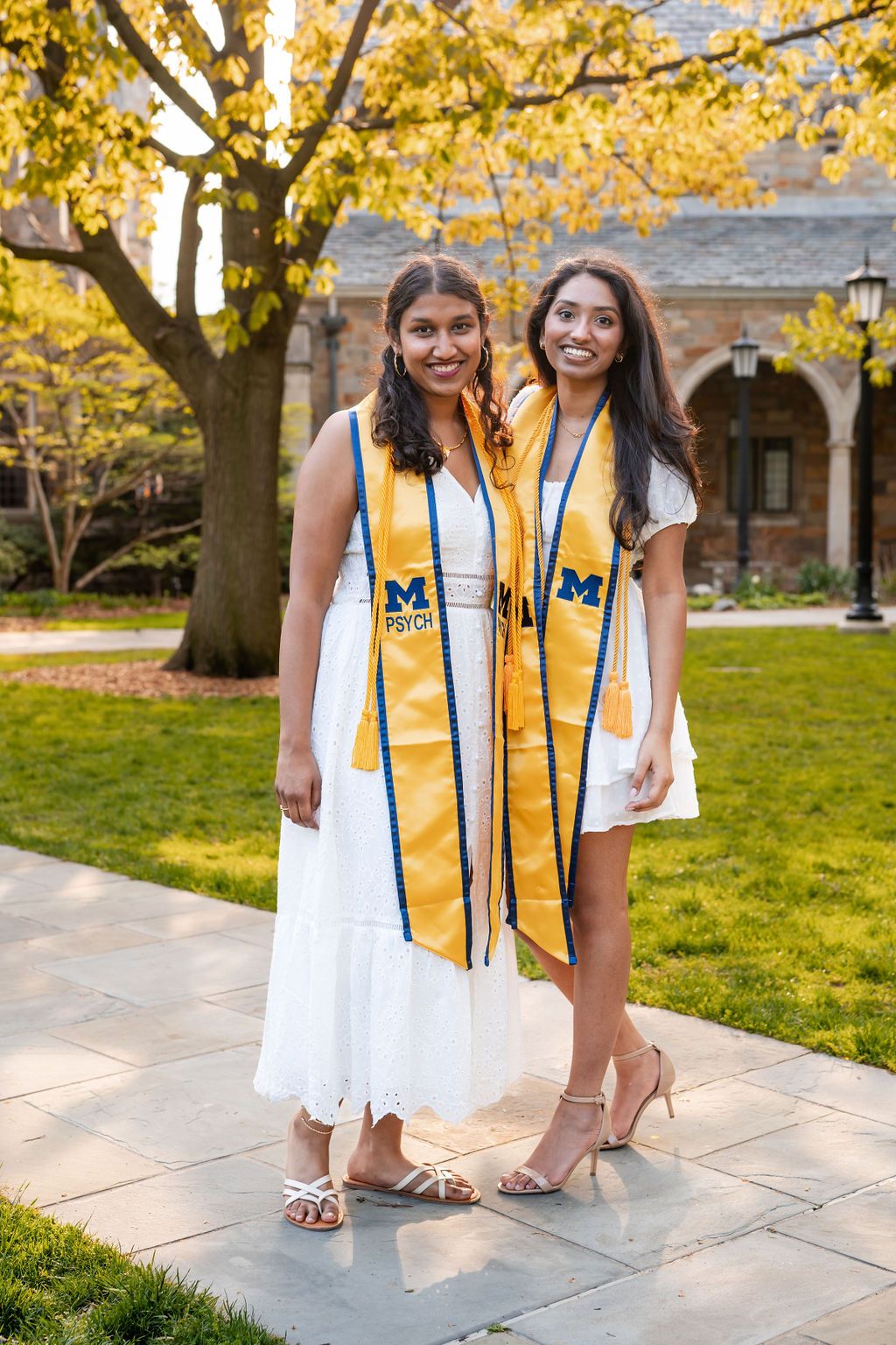 Two graduates in white dresses and yellow stoles pose together on a university campus with fall foliage in background.