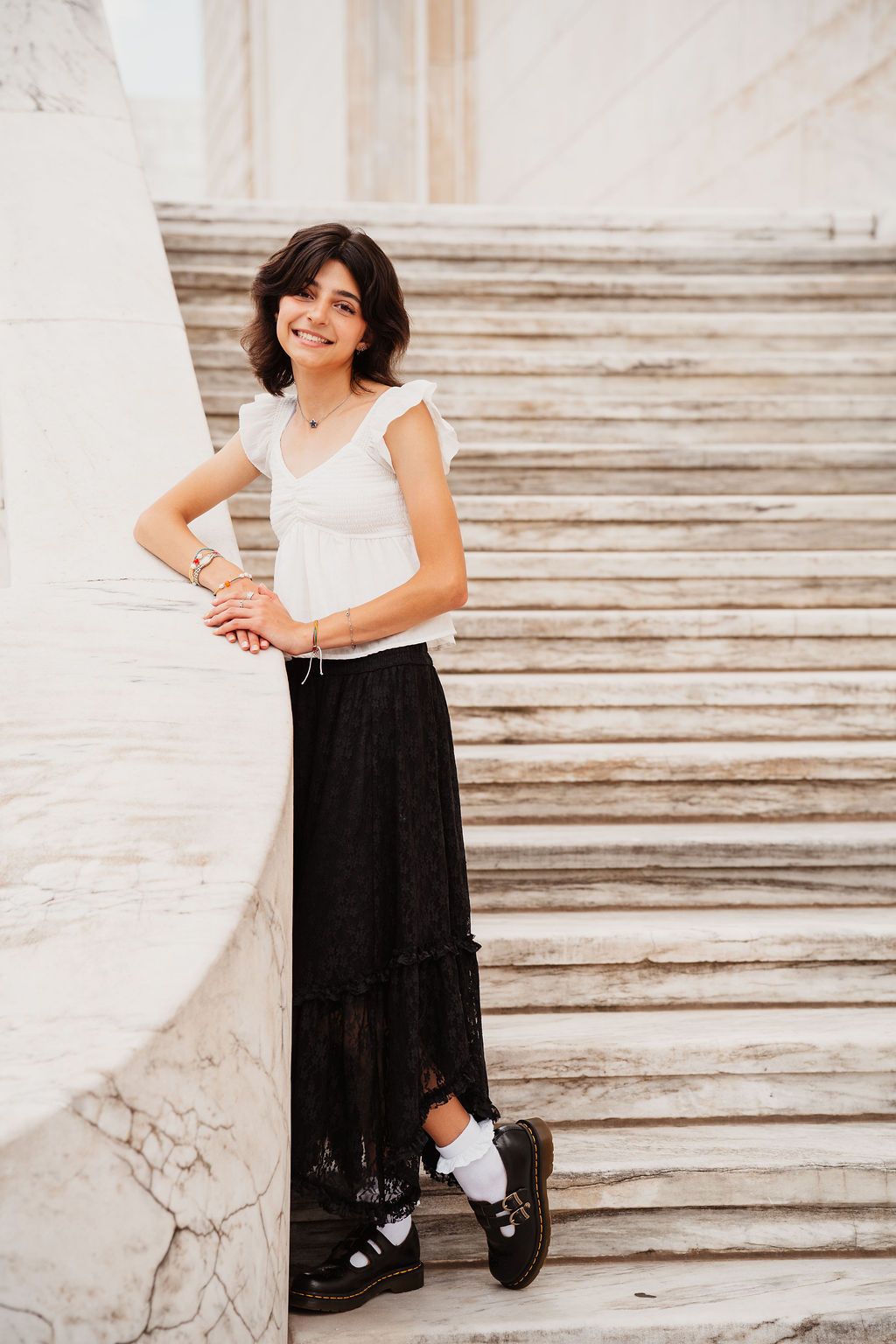 Young woman in white top and black maxi skirt smiling on marble steps.