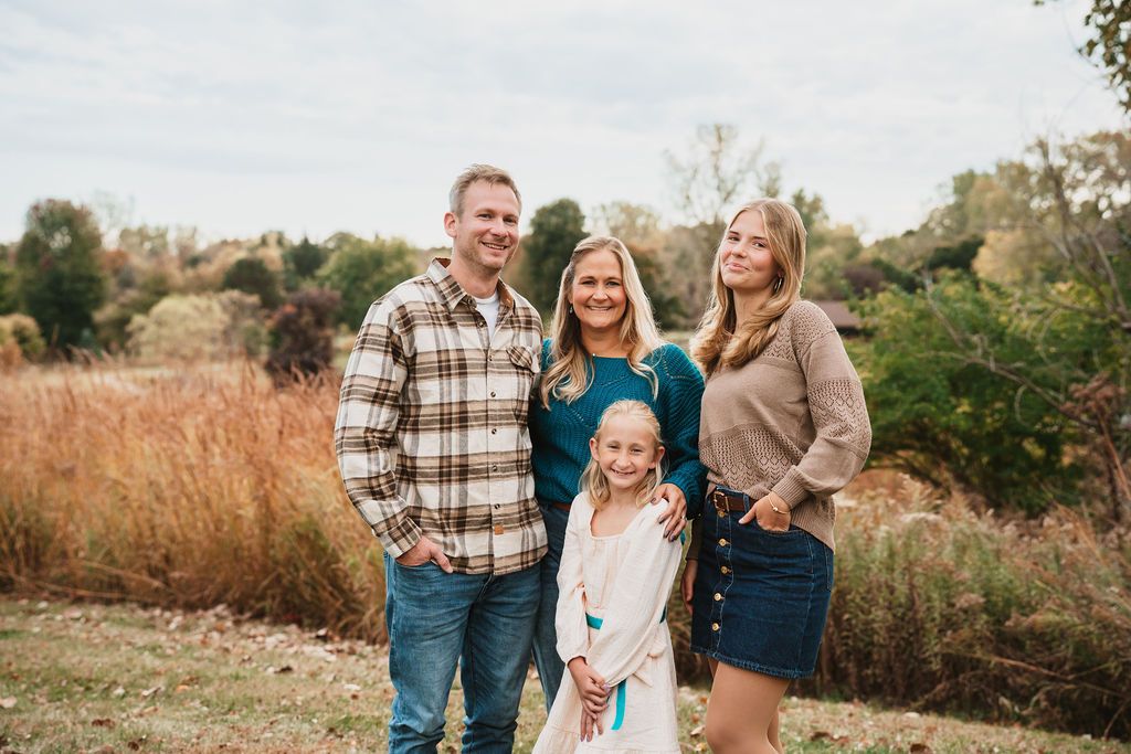 Family of four smiling outdoors in autumn, wearing casual fall attire amid golden grasses and trees.