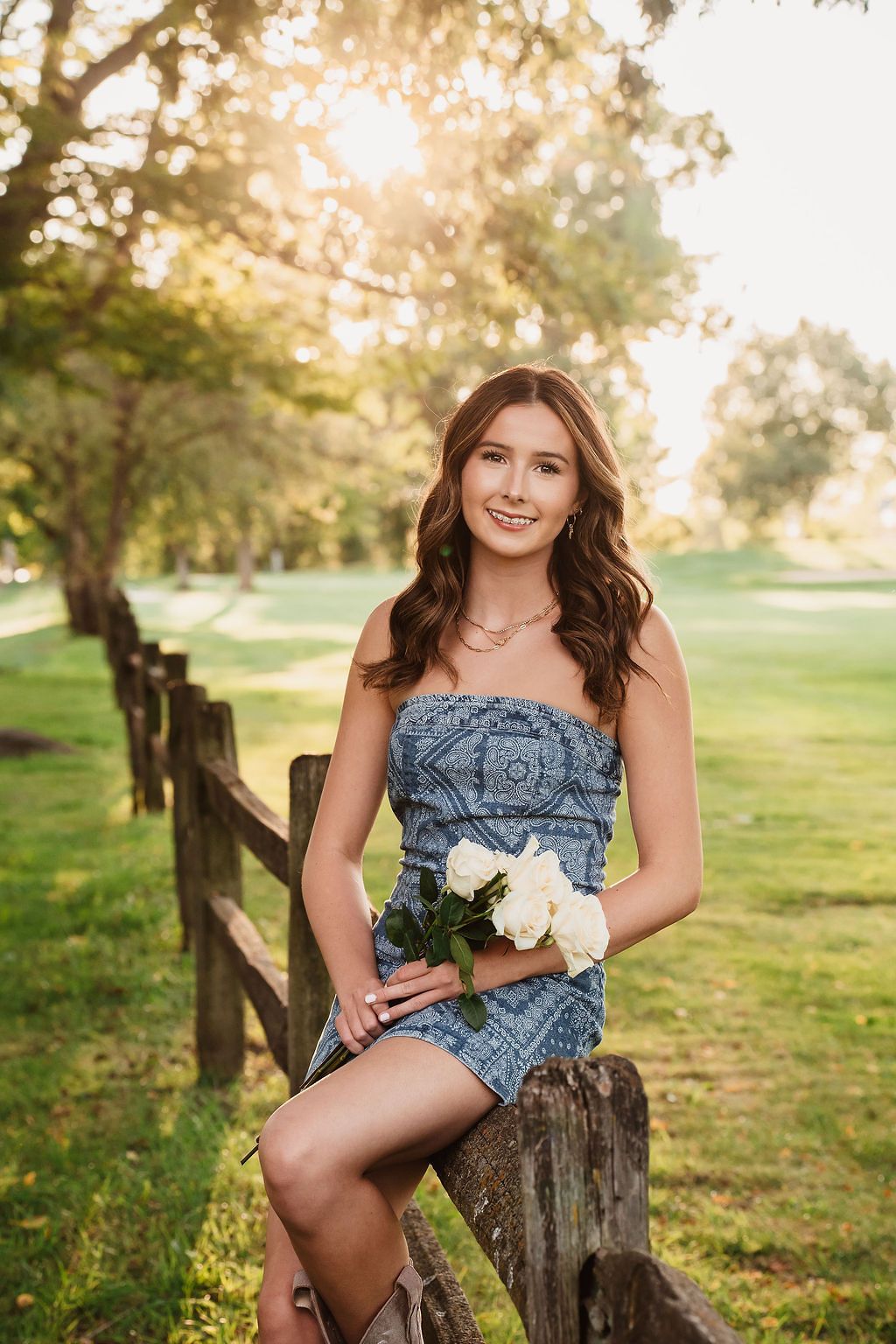 Young woman in denim dress holding white flowers sits on rustic fence in golden sunlight park setting.
