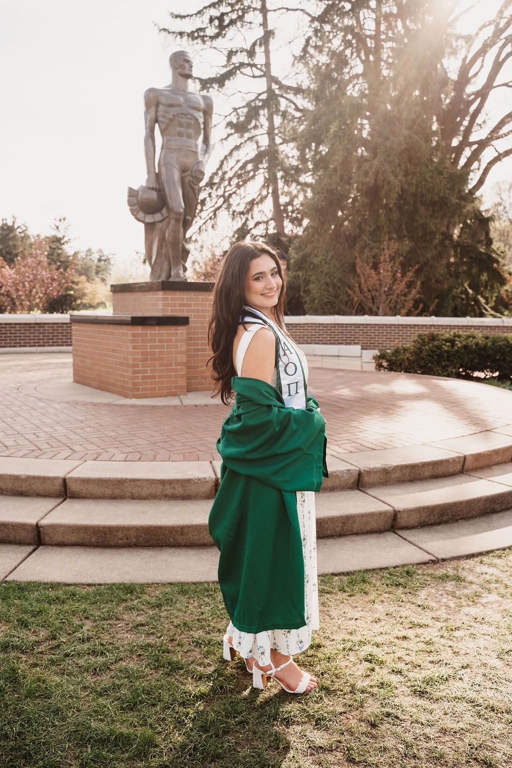 Young woman in green graduation gown poses in front of Spartan statue at Michigan State University.