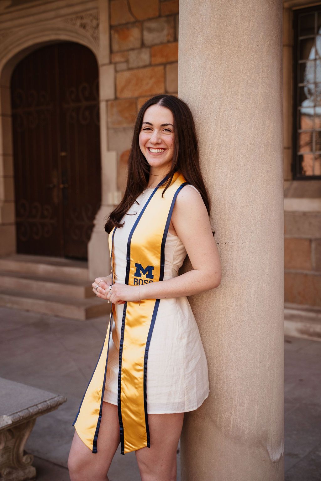 Smiling young woman in a white dress wearing a University of Michigan graduation stole, leaning against a stone column.