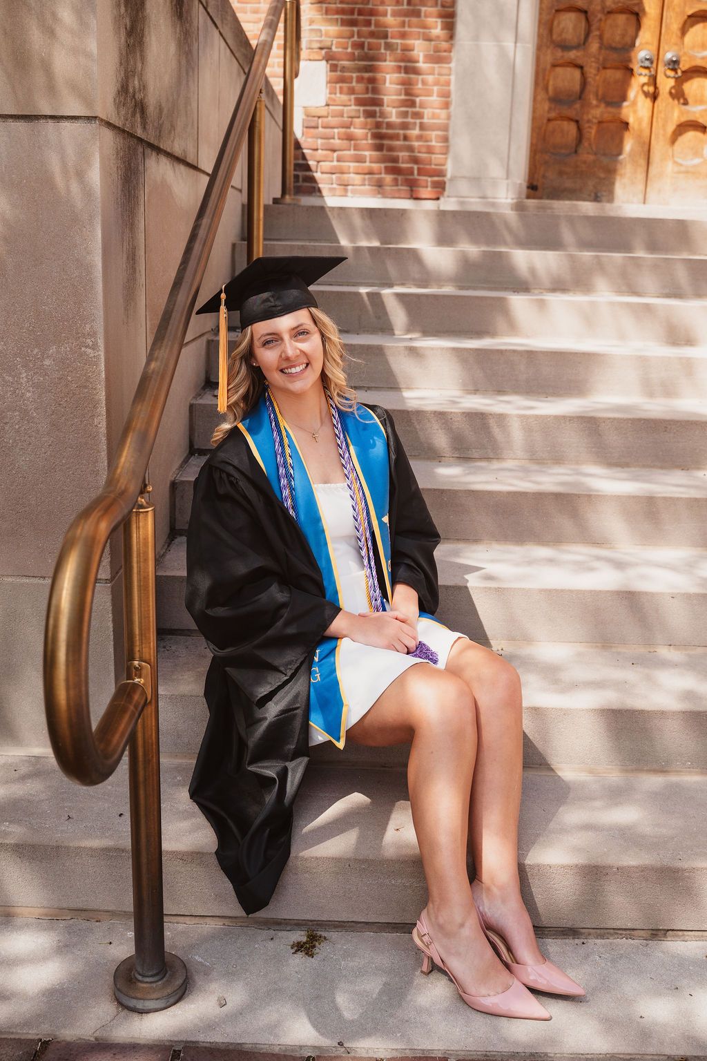Smiling female graduate in black cap and gown with blue sash sitting on stone steps holding diploma.