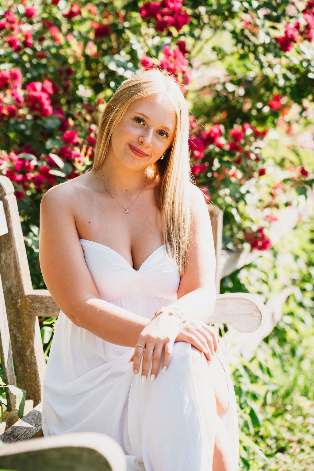 Blonde woman in white strapless dress sitting in garden chair with red flowering bushes in background.