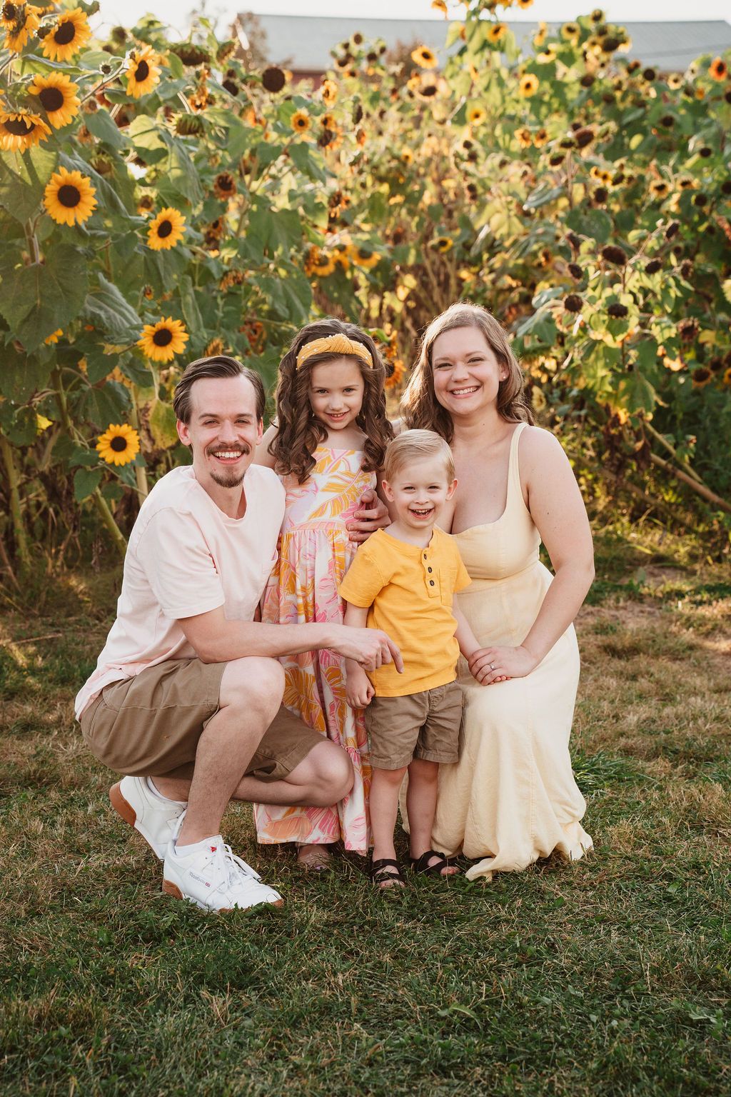 Happy family of four posing in a sunflower field, parents smiling with two young children in yellow outfits.