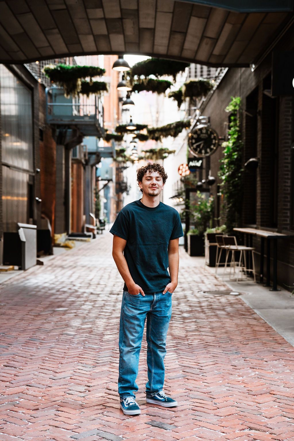 Young man in blue jeans and black t-shirt standing on brick alley with hanging plants and string lights.