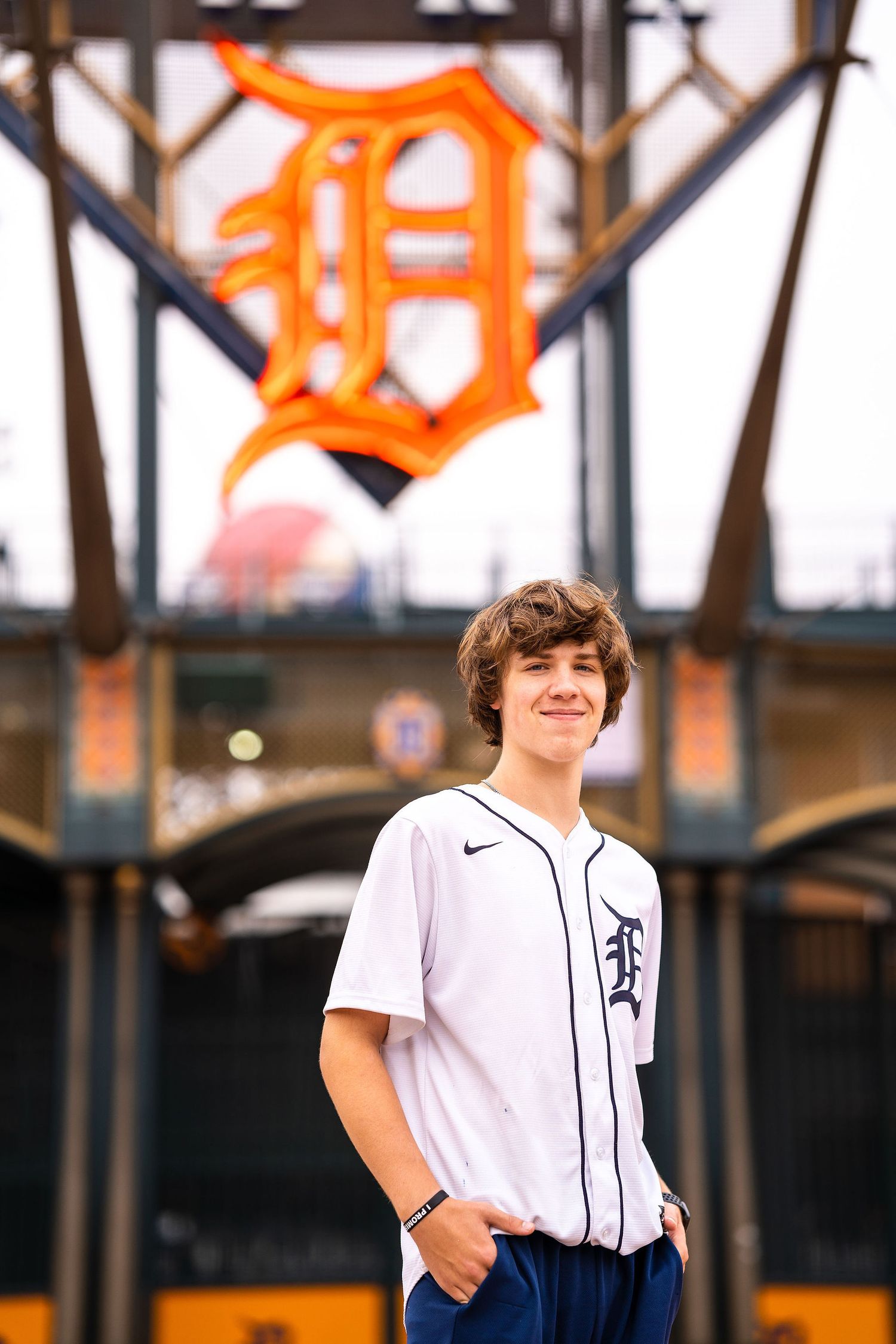 Teen boy in Detroit Tigers jersey smiles at Comerica Park with iconic Old English D logo in background.