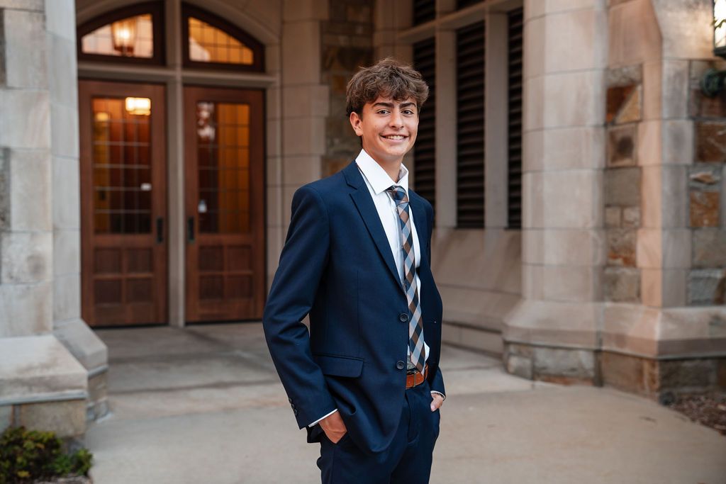 Young man in navy blue suit and striped tie posing confidently outside a stone building.