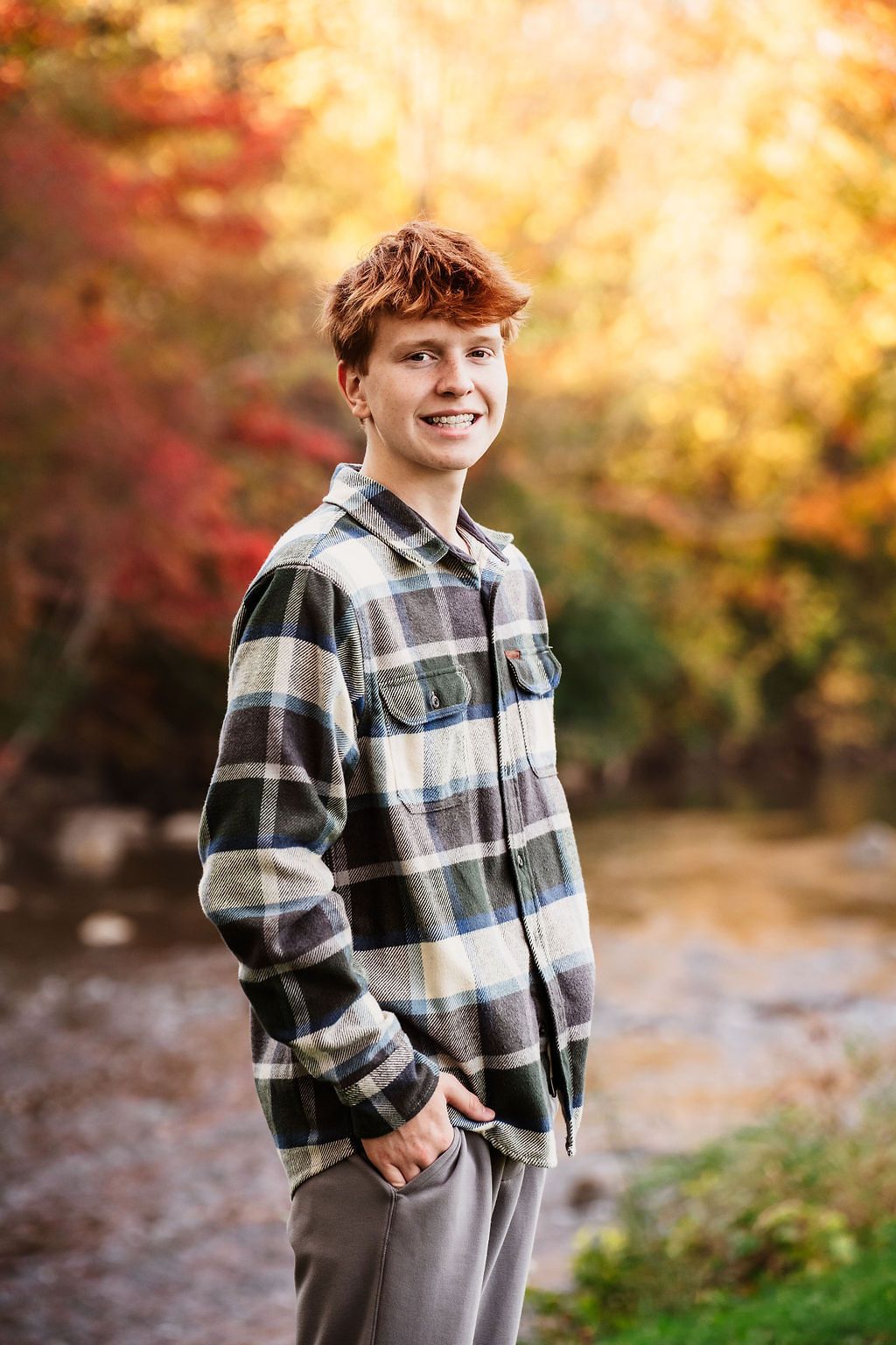 Smiling teenage boy with red hair in plaid shirt standing outdoors amid vibrant autumn foliage near a stream.