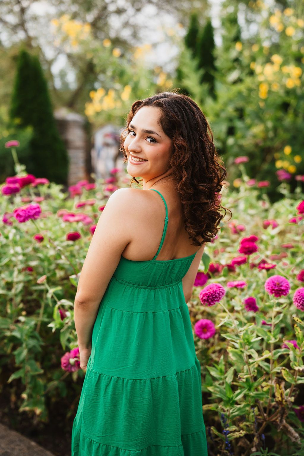 A woman in an emerald green sundress poses in a garden filled with vibrant pink flowers and yellow blooms.