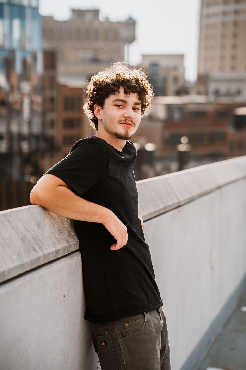 Person in black t-shirt leaning against concrete wall in urban setting with buildings visible in background.