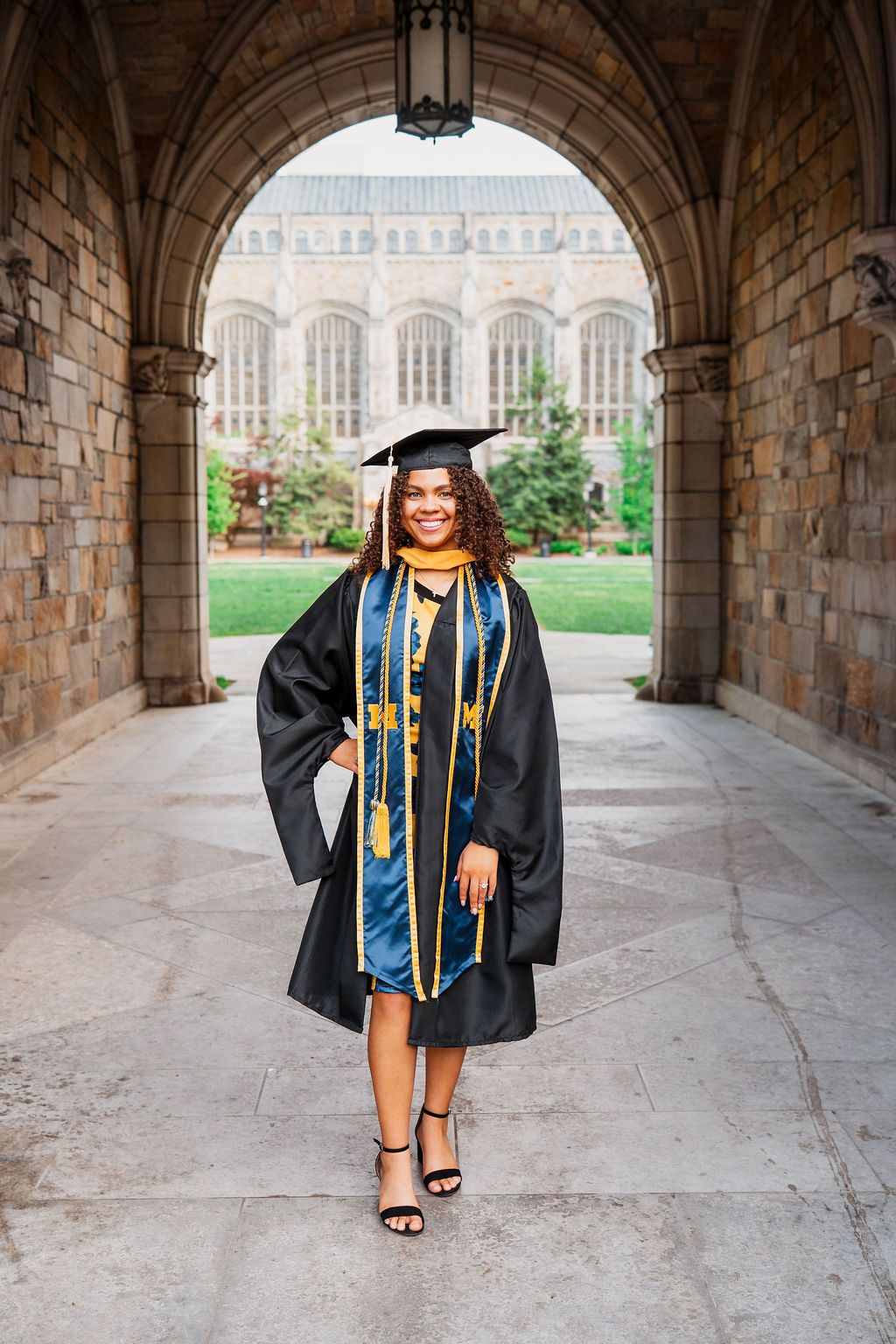 Graduate in cap and gown stands under stone archway on university campus with Gothic architecture in background.