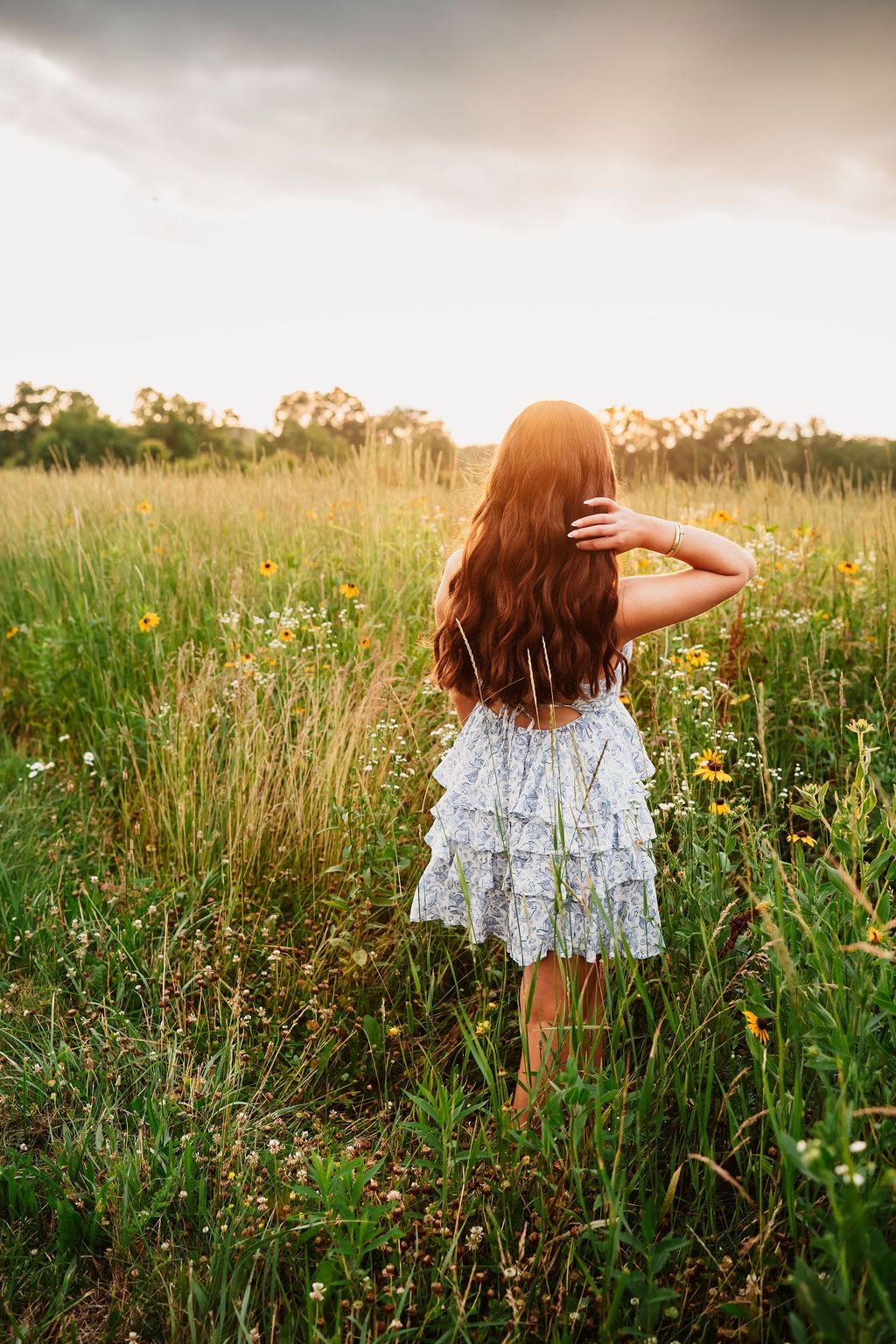 A person with long red hair wearing a white ruffled dress stands in a wildflower meadow at sunset.