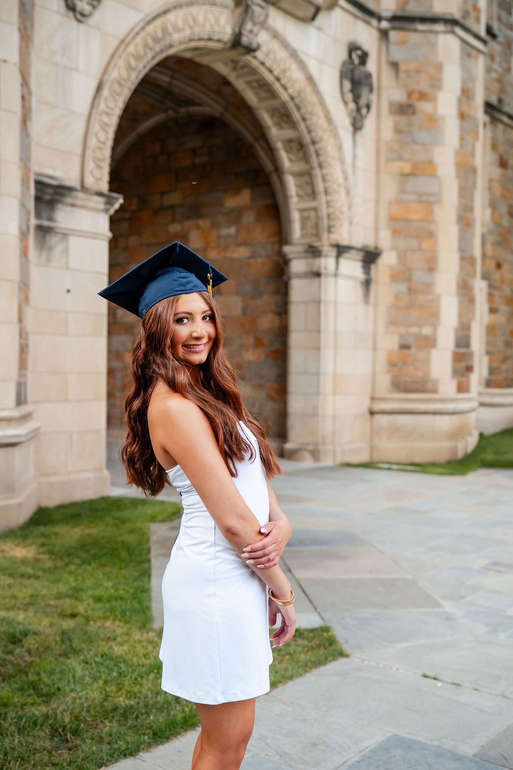 Graduate in white dress and cap poses for photos in front of historic stone archways on campus.