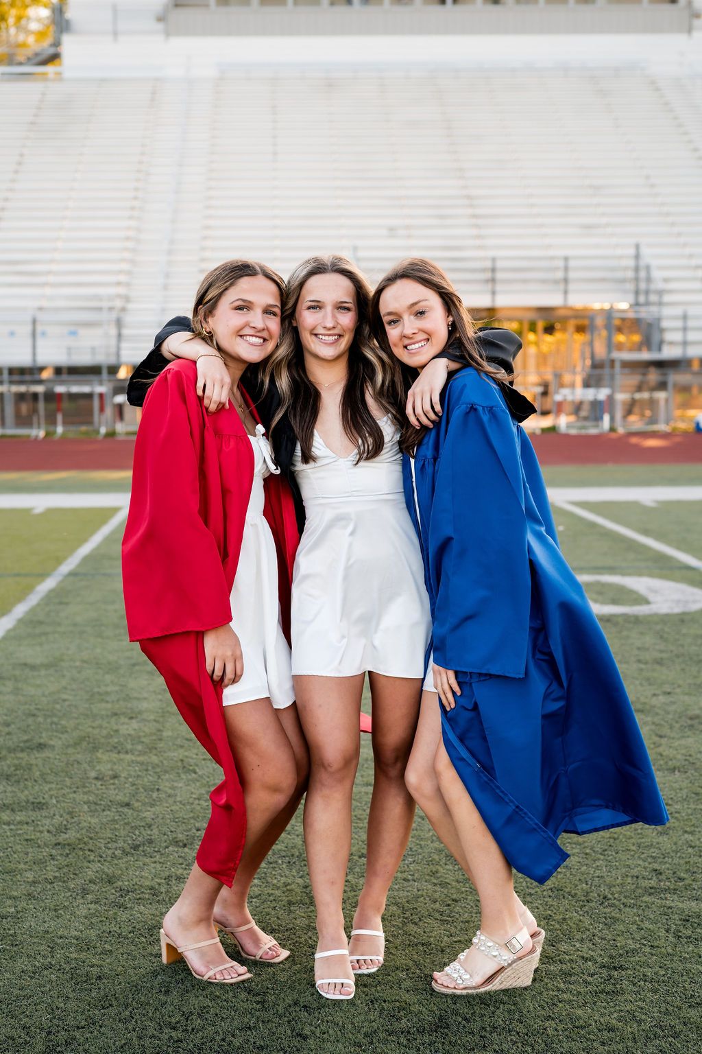 Friends in red and blue graduation attire smile together on an athletic field.