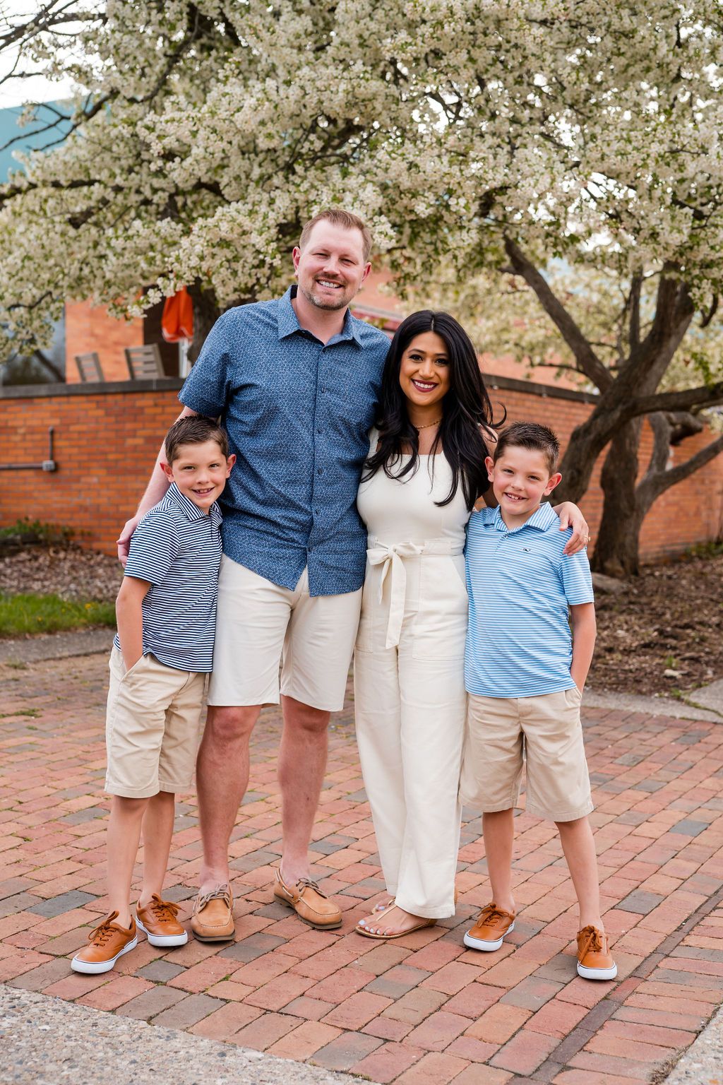 A family of four stands together on brick pavement under a blooming spring tree.