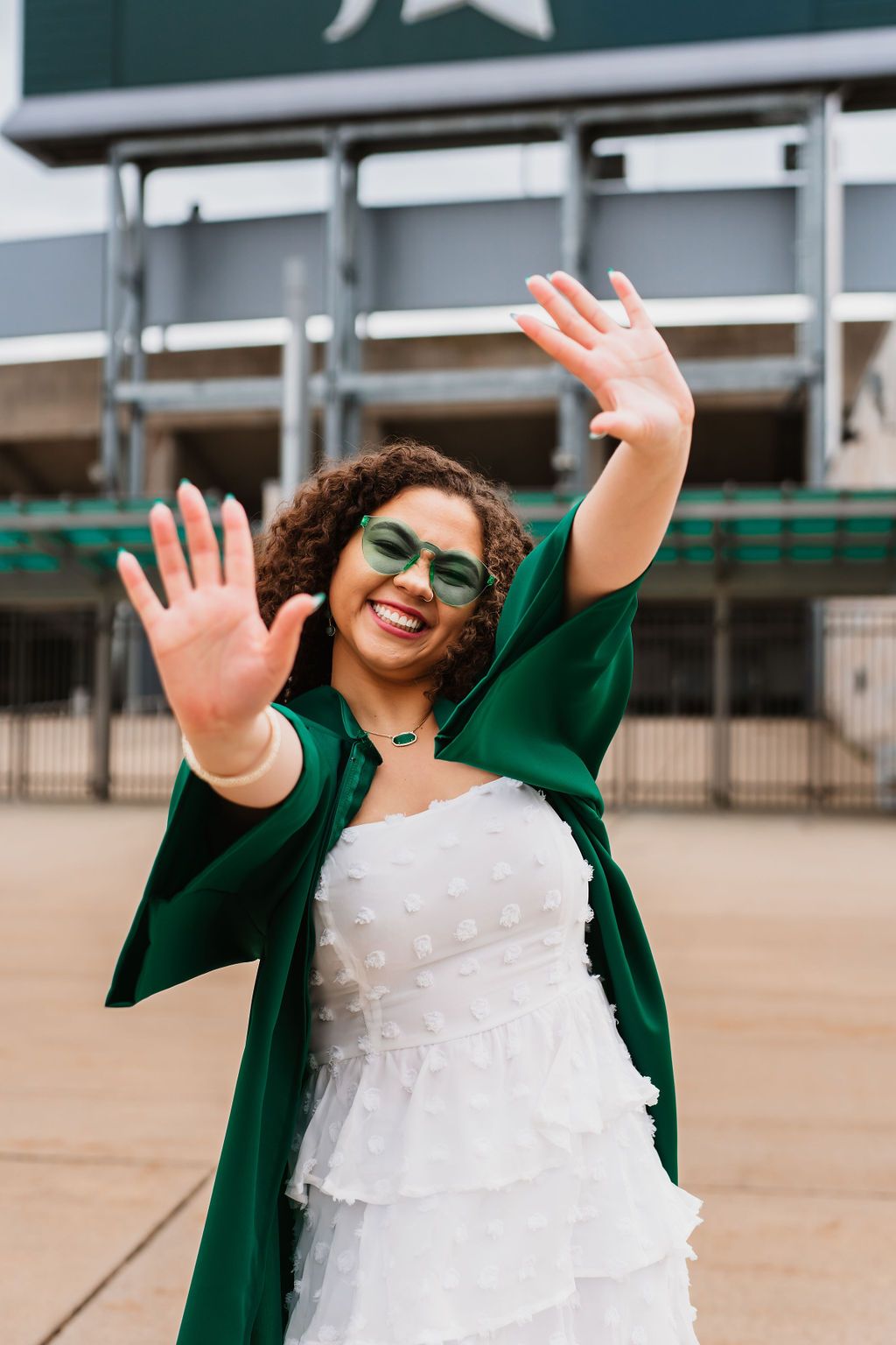Person in white dress and green jacket throws arms up joyfully outside a stadium building.