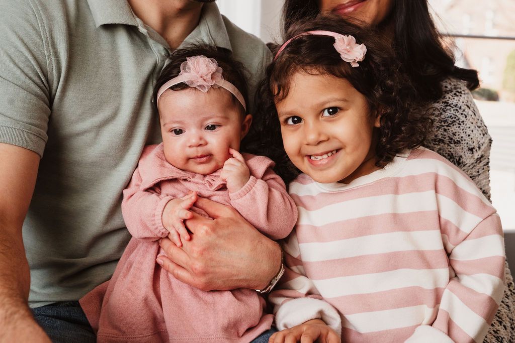 A family portrait featuring a man and woman holding a smiling toddler girl and a baby in pink outfits.