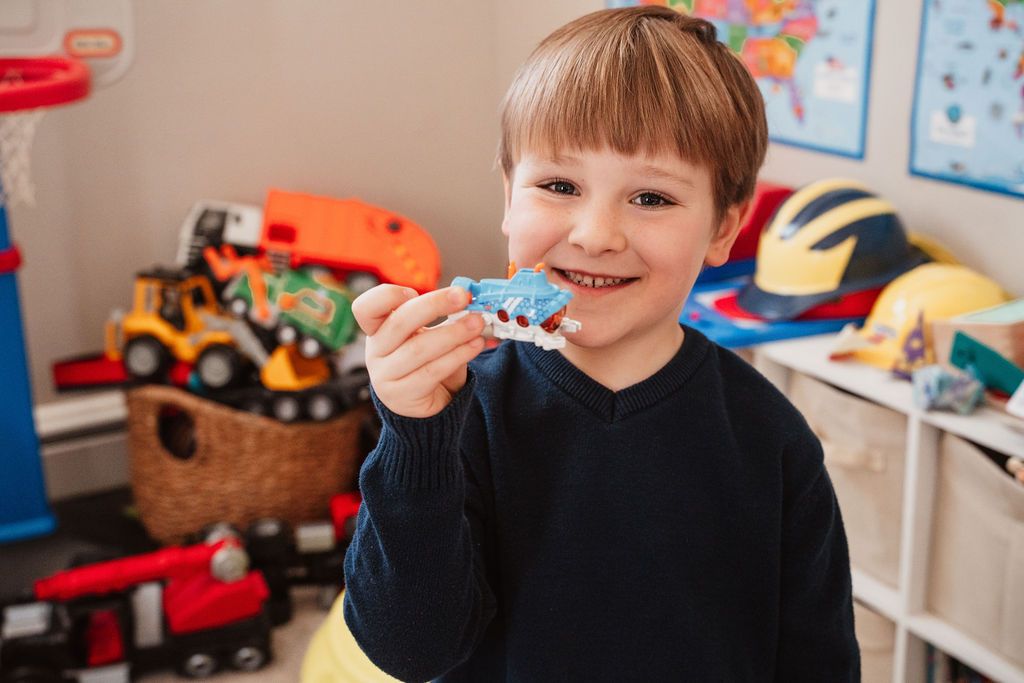 Smiling young boy holding a small blue toy in a colorful playroom filled with trucks and toys.