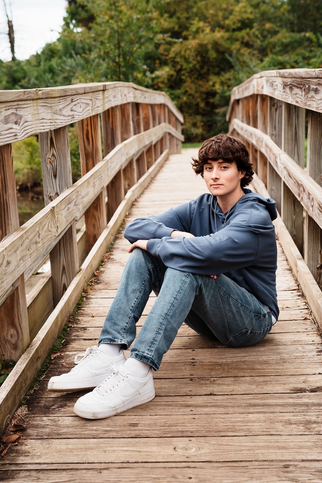 Teen boy in blue hoodie sitting on wooden bridge deck surrounded by trees.
