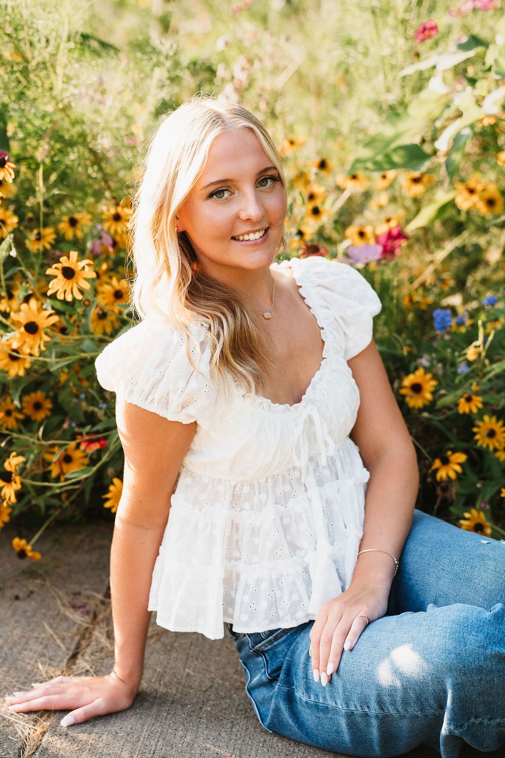 Smiling blonde woman in white ruffled top and jeans sitting outdoors among vibrant yellow wildflowers.