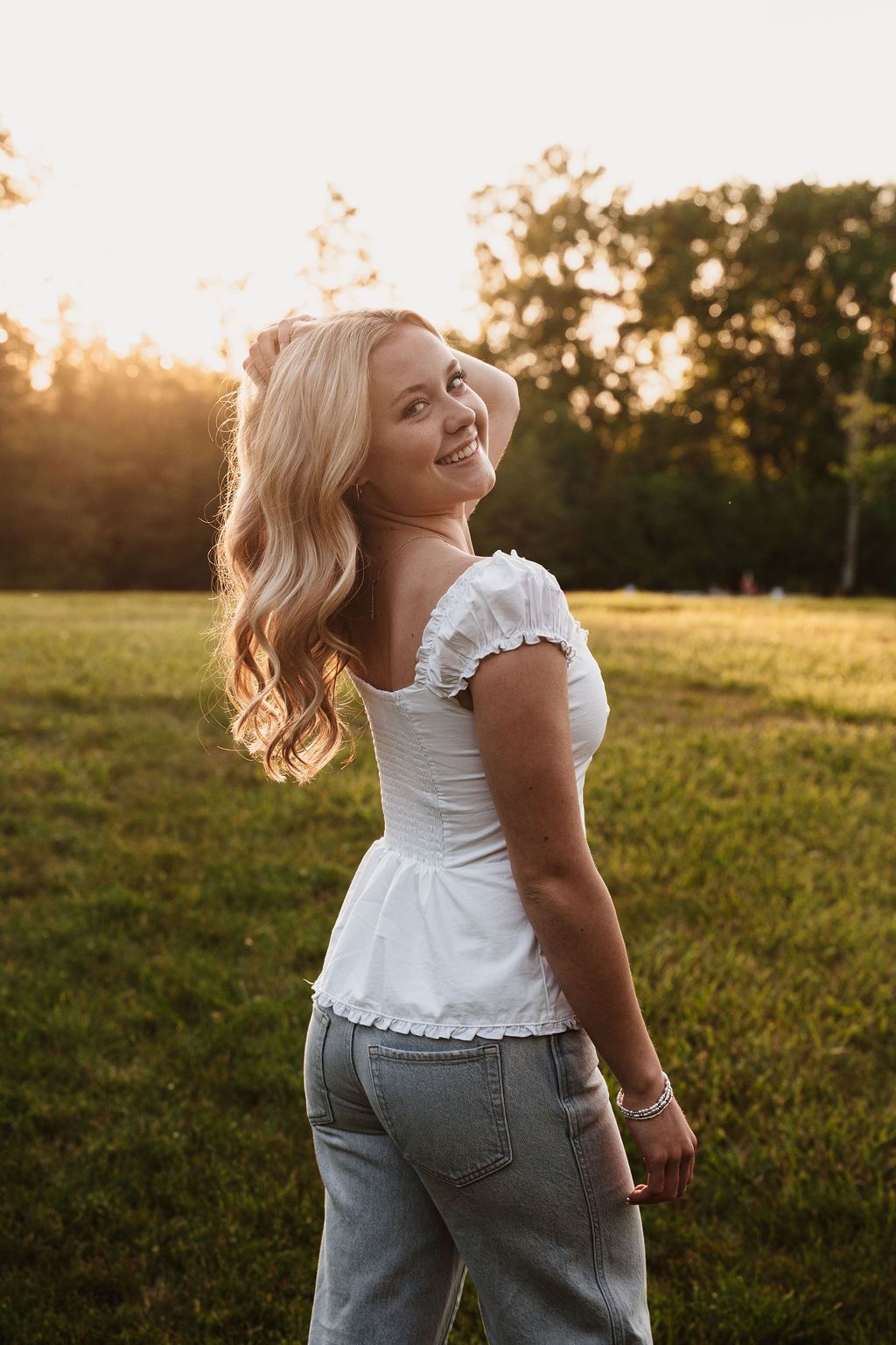 Smiling blonde woman in white top and jeans poses in sunlit green field at golden hour.