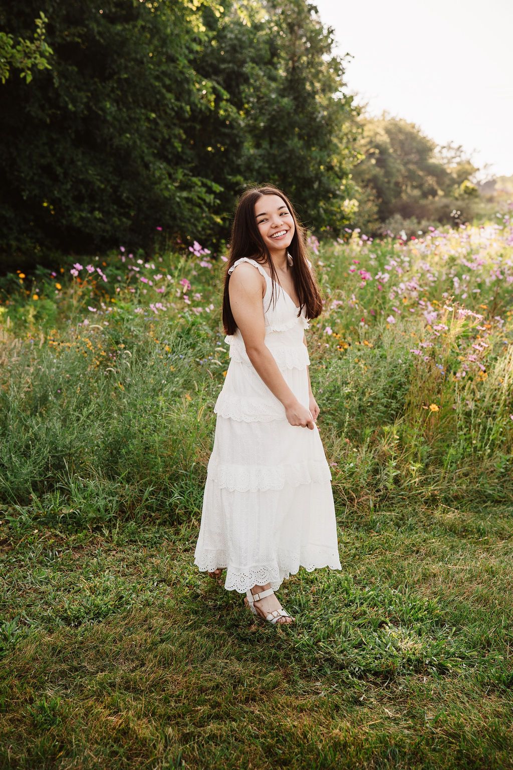 Young woman in white midi dress smiling in a wildflower meadow with lush green trees in golden sunlight.