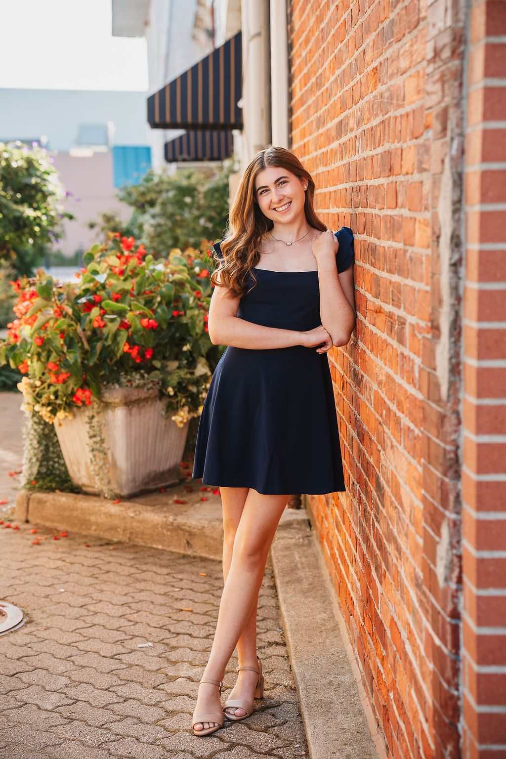 Young woman in navy mini dress leaning against brick wall, smiling, with red flowers in background.