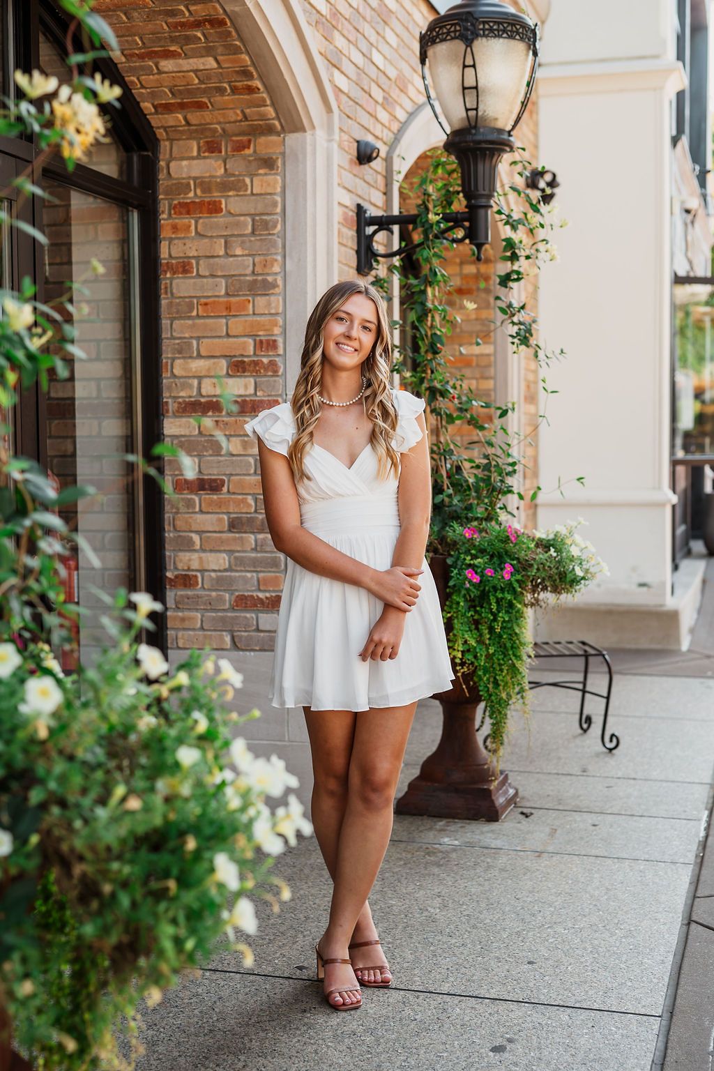 Smiling young woman in white mini dress standing on brick sidewalk near lamp post and flowers.